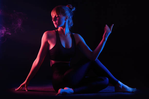 Workout under neon light. Slender young woman yogi doing yoga asana Marichyasana. Low key yoga studio photosession. Black background, red and blue color light. Incense smoke, a meditative space. stock photo