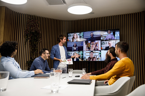 Candid shot of small group of businessmen sitting around a conference table and discussing business with their coworkers on the flat screen over a video conference.