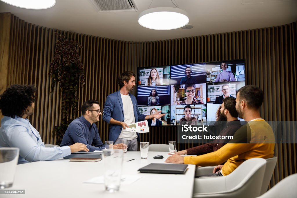 Group of businessmen having a video conference meeting with their coworkers Candid shot of small group of businessmen sitting around a conference table and discussing business with their coworkers on the flat screen over a video conference. Meeting Stock Photo