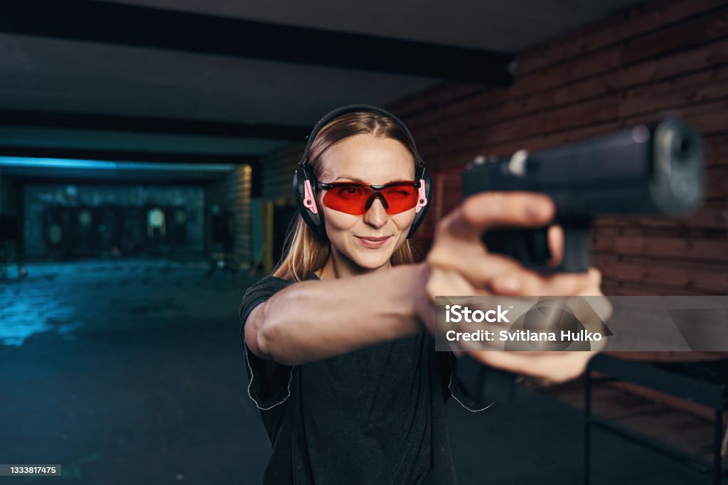 Young woman concentrated on shooting at a target Waist-up portrait of a focused female sniper in safety glasses and earmuffs firing a pistol with both hands Target Shooting Stock Photo