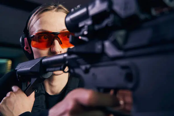 Serious focused young Caucasian woman in protective eyewear shooting a modern weapon with a riflescope