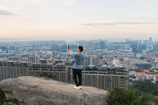 Asian man takes a selfie on the rock mountain after climbing, with city buildings in the background