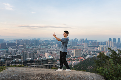 A man using a smart phone to take a photo, the background is on the rock after climbing, facing the city buildings