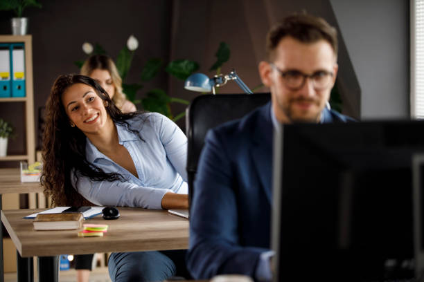 Peeking at her office crush during work Selective focus shot of cheerful young businesswoman sitting at her desk and peeking at her male colleague, an office crush. work romance stock pictures, royalty-free photos & images