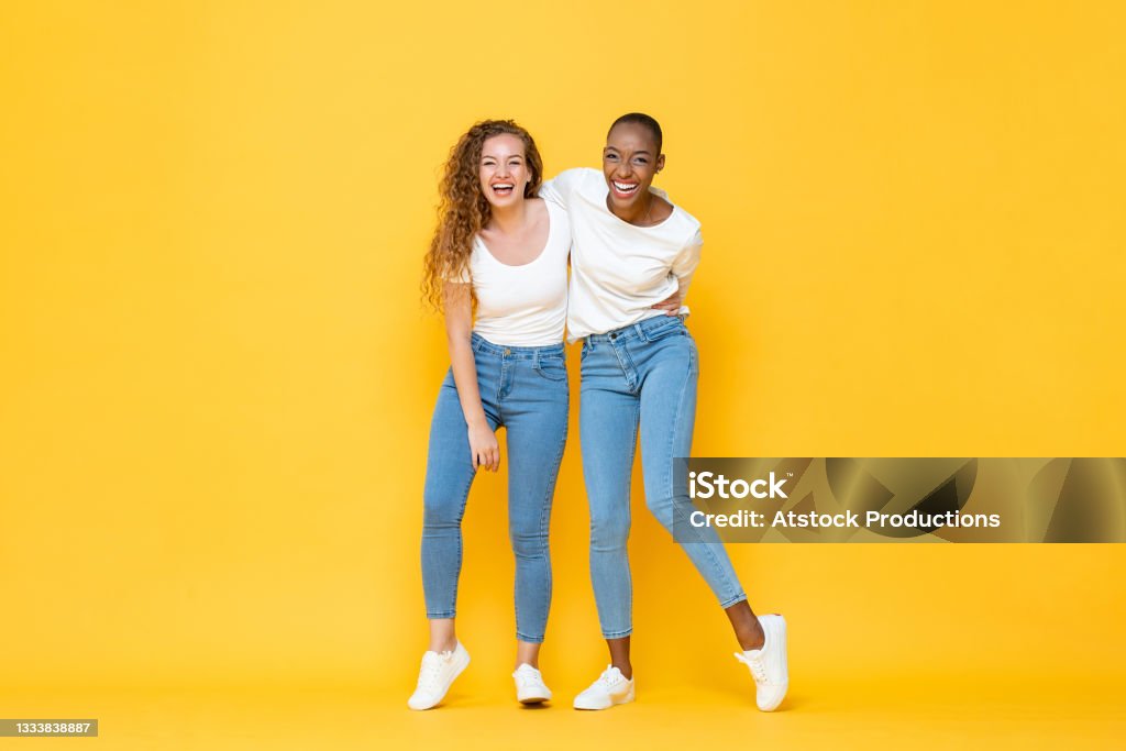 Happy two interracial woman friends smiling and holding each other in isolated studio yellow color background Friendship Stock Photo