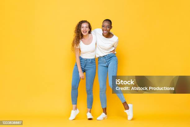 Happy Two Interracial Woman Friends Smiling And Holding Each Other In Isolated Studio Yellow Color Background Stock Photo - Download Image Now