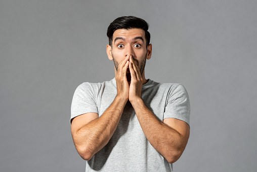 Shocked young Caucasian man with hands covering mouth in isolated light gray studio background