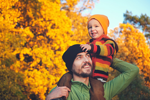 Cute little child boy sitting on his father's shoulders on background of beautiful autumn forest. Time together dad and son outdoor in fall season.