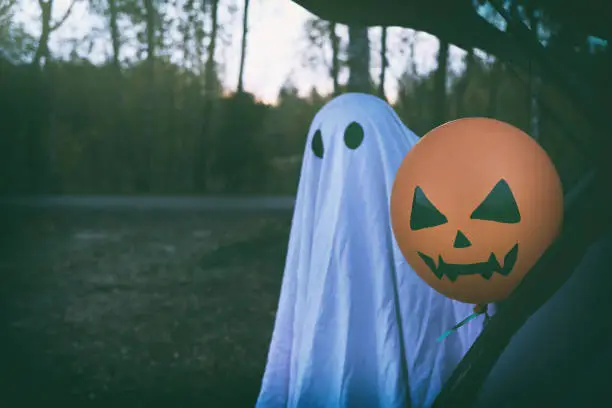 Halloween concept. Cute little child in ghost costume holding scary face orange air balloon while sitting inside car trunk near twilight autumn forest road.