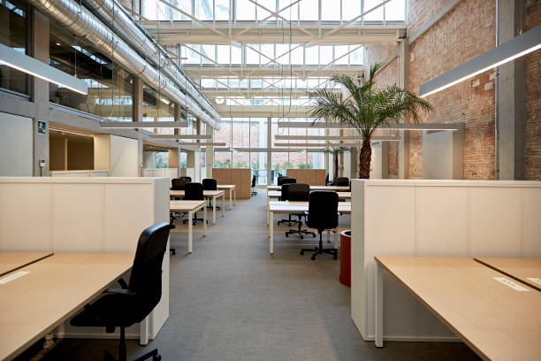 Open Plan Industrial-Style Office Space Wide angle view of unoccupied desks and chairs on ground floor of renovated building with exposed brick wall, high ceiling, and visible second floor windows. commercial real estate stock pictures, royalty-free photos & images