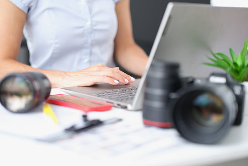 Woman typing on laptop keyboard at table with camera lenses closeup. Content creation concept