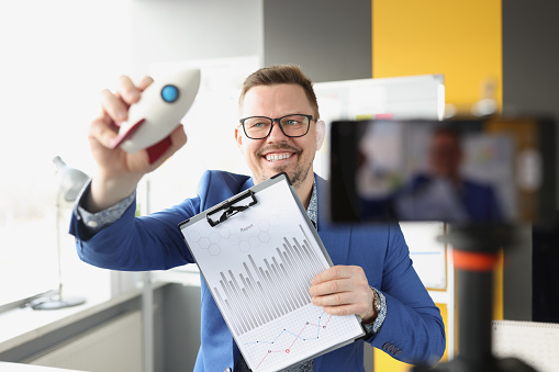 Smiling male coach holding toy rocket and clipboard with documents in front of mobile phone camera. Conducting online courses on starting business development concept