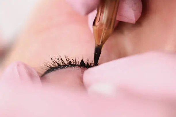 Photo of Master making permanent make-up of space between eyelashes to woman client in beauty salon closeup