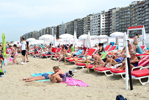 Blankenberge, Belgium - August 12, 2021: Wealthy tourists lying in a row can afford to sunbathe on red beach chairs. Beautiful terraced apartments behind white beach umbrellas.