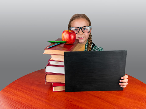 Little girl in glasses with book stack and red apple on desk working in class. Back to School knowledges education concept. Cute smiling Primary school student caucasian child portrait in classroom.
