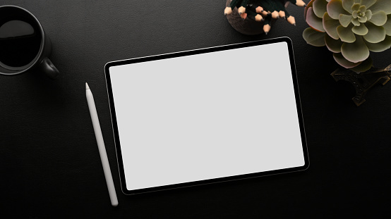 Portable tablet white screen mockup with stylus pen on dark working space with house plant, pencils, coffee, black background, overhead