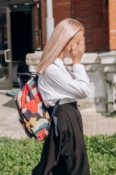 A sad schoolgirl in a uniform and with a backpack is walking out of school, covering her face with her hands.School concept.