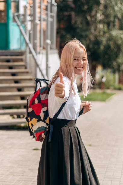 A positive blonde teenage girl in a school uniform and a backpack is walking down the street to school and showing a thumbs up gesture.Back to school concept A positive blonde teenage girl in a school uniform and a backpack is walking down the street to school and showing a thumbs up gesture.Back to school concept. 15 year old blonde girl stock pictures, royalty-free photos & images