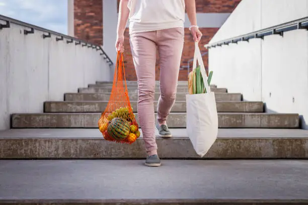 Woman walking at stairs and carrying reusable mesh bag after shopping groceries in city. Sustainable lifestyle with zero waste and plastic free