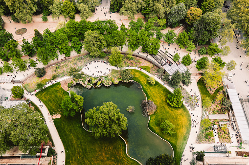 View of verdant gardens from top of Eiffel Tower. Pathways surrounded by lush greenery, wooded area, basin. Famous Champ de Mars in Paris, France. Walking people in historical place.