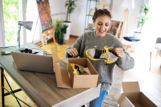 Photo of Young woman unpacking boots from cardboard box that she have received after online shopping
