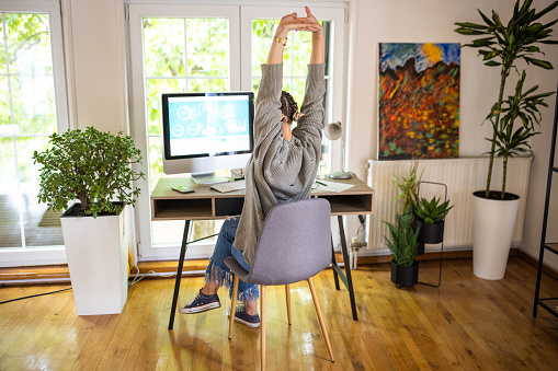 Young woman sitting at desk and stretching while telecommuting, using computer from home
