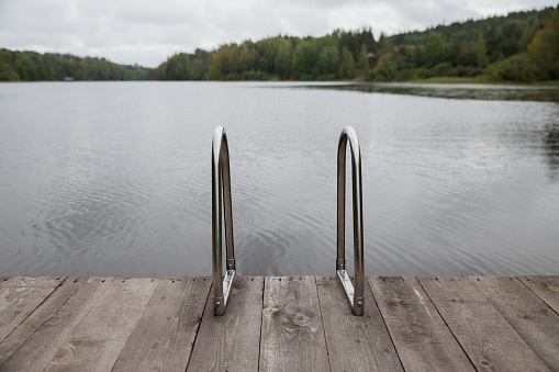 Metal handrail for descent into the lake, iron ladder for swimmers. Wooden pier on the lake, cloudy sky and calm lake water on background.