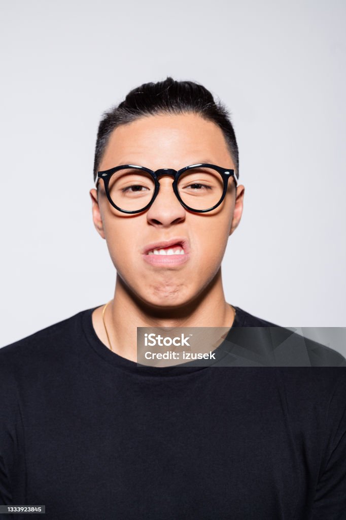 Angry asian young man in black clothes Headshot of displeased asian young man wearing black t-shirt and eyeglasses, looking at camera. Studio shot, grey background. Human Face Stock Photo