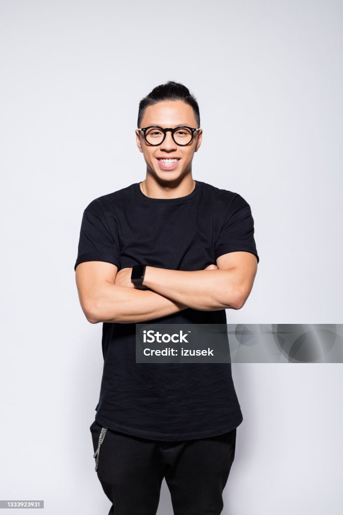Cheerful asian young man in black clothes Portrait of handsome asian young man wearing black t-shirt, pants and eyeglasses, standing with arms crossed and laughing at camera. Studio shot, grey background. Arms Crossed Stock Photo