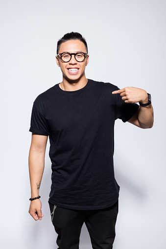 Portrait of handsome asian young man wearing black t-shirt, pants and eyeglasses, pointing with index finger at his chest and laughing at camera. Studio shot, grey background.