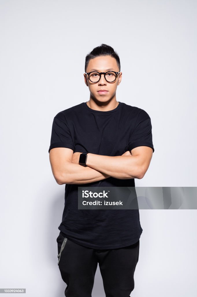 Confident asian young man in black clothes Portrait of handsome asian young man wearing black t-shirt, pants and eyeglasses, standing with arms crossed and looking at camera. Studio shot, grey background. Portrait Stock Photo
