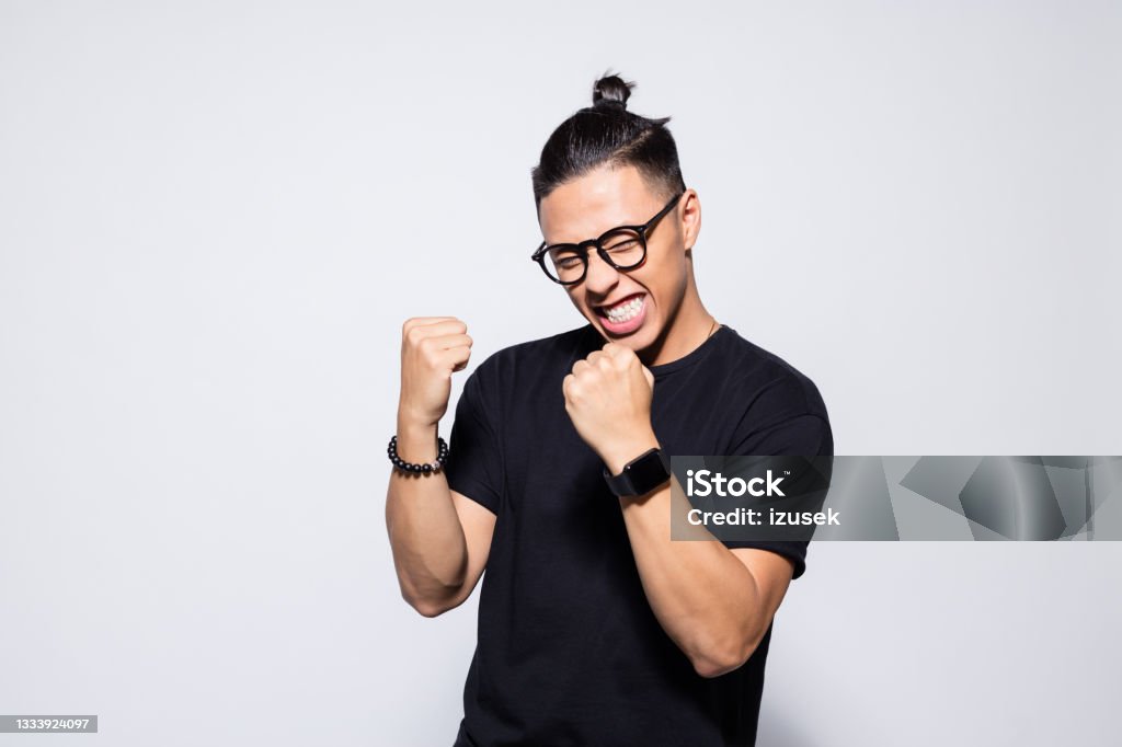 Excited asian young man in black clothes Portrait of happy asian young man wearing black t-shirt and eyeglasses, clenching fists. Studio shot, grey background. Men Stock Photo