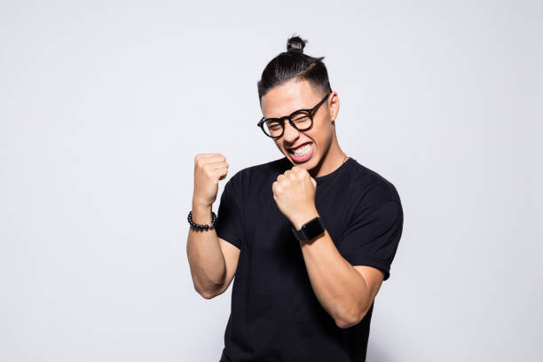 Excited asian young man in black clothes Portrait of happy asian young man wearing black t-shirt and eyeglasses, clenching fists. Studio shot, grey background. black nerd stock pictures, royalty-free photos & images