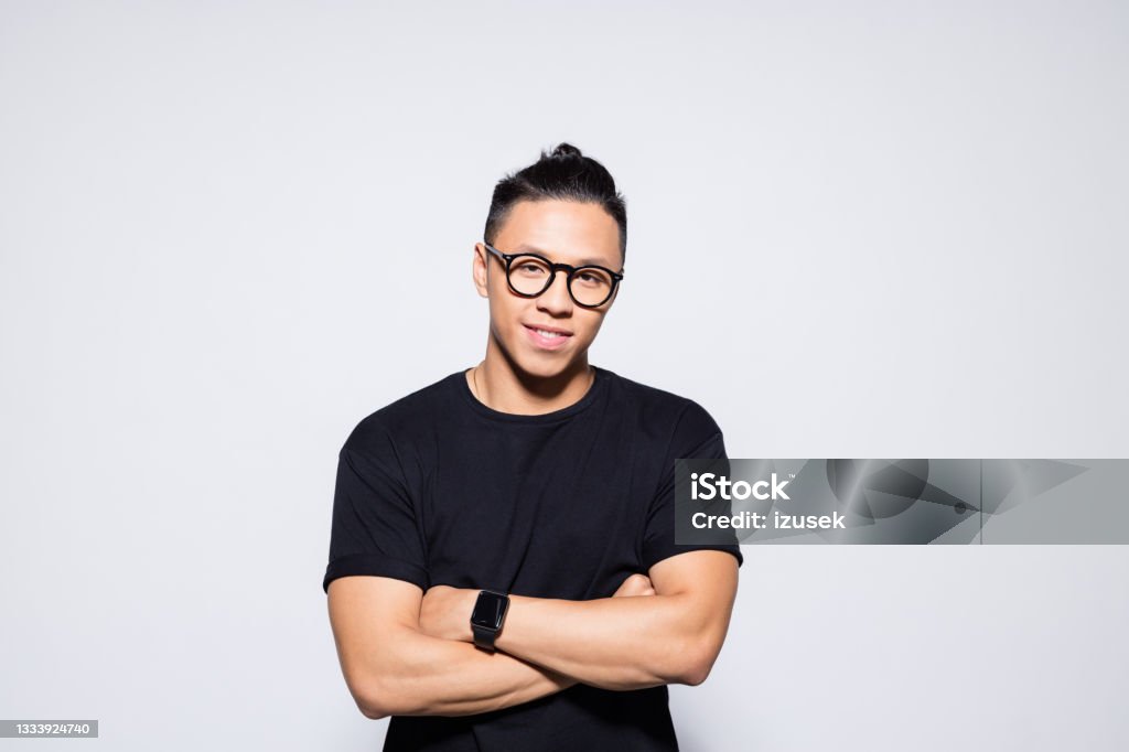 Friendly asian young man in black clothes Portrait of handsome asian young man wearing black t-shirt and eyeglasses, standing with arms crossed and smiling at camera. Studio shot, grey background. Asian and Indian Ethnicities Stock Photo