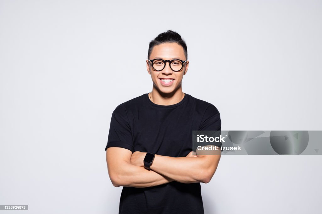 Friendly asian young man in black clothes Portrait of handsome asian young man wearing black t-shirt and eyeglasses, standing with arms crossed and smiling at camera. Studio shot, grey background. Men Stock Photo