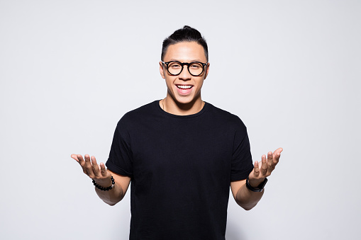 Portrait of handsome asian young man wearing black t-shirt and eyeglasses, smiling at camera with raised hand. Studio shot, grey background.