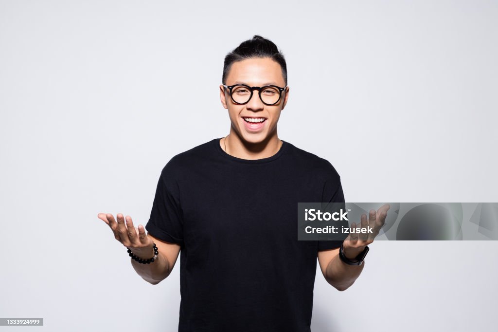 Happy asian young man in black clothes Portrait of handsome asian young man wearing black t-shirt and eyeglasses, smiling at camera with raised hand. Studio shot, grey background. Men Stock Photo