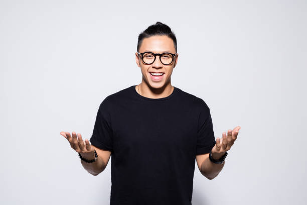 Happy asian young man in black clothes Portrait of handsome asian young man wearing black t-shirt and eyeglasses, smiling at camera with raised hand. Studio shot, grey background. black nerd stock pictures, royalty-free photos & images