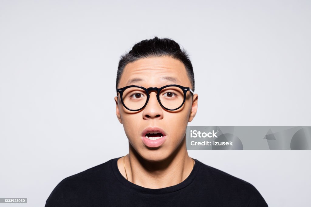 Shocked asian young man in black clothes Headshot of terrified asian young man wearing black t-shirt and eyeglasses, looking at camera. Studio shot, grey background. Shock Stock Photo