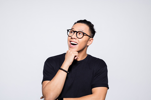 Portrait of excited asian young man wearing black t-shirt and eyeglasses, looking away with hand on chin. Studio shot, grey background.