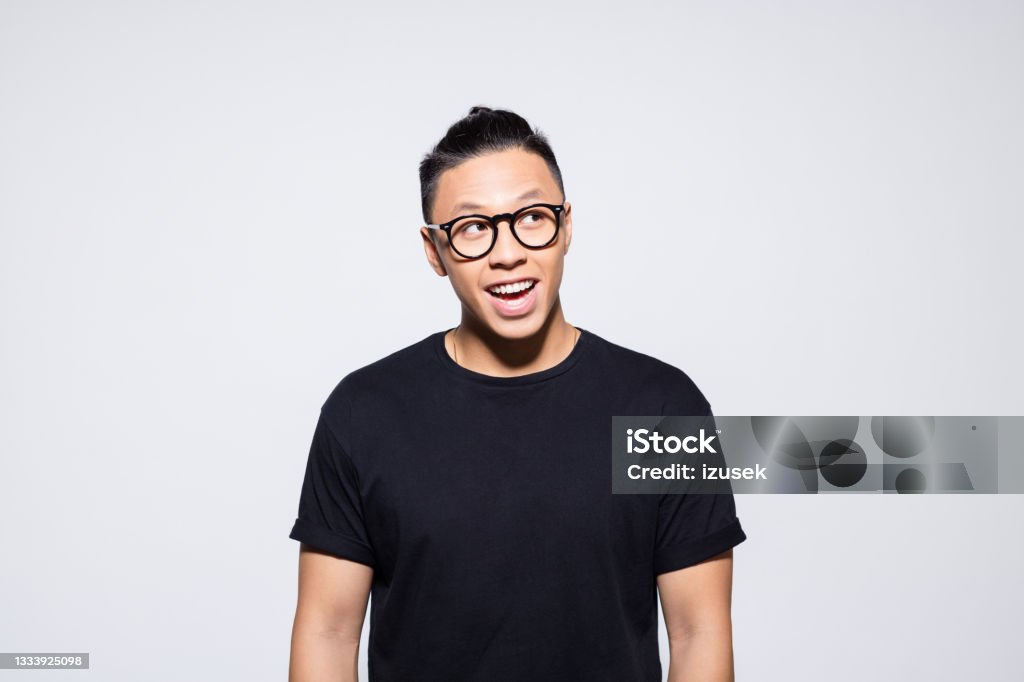 Surprised asian young man in black clothes Portrait of cheerful asian young man wearing black t-shirt and eyeglasses, looking away. Studio shot, grey background. Portrait Stock Photo