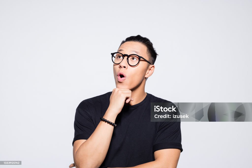 Surprised asian young man in black clothes Portrait of cheerful asian young man wearing black t-shirt and eyeglasses, looking away with hand on chin. Studio shot, grey background. Adult Stock Photo