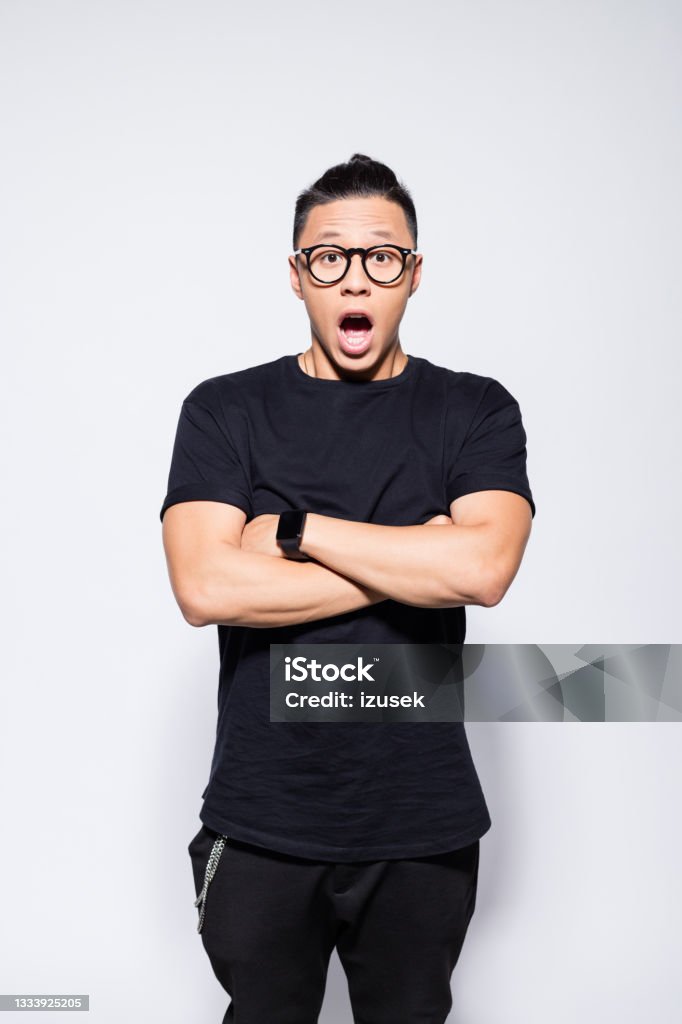 Surprised asian young man in black clothes Portrait of handsome asian young man wearing black t-shirt, pants and eyeglasses, standing with arms crossed and looking at camera with mouth open. Studio shot, grey background. Arms Crossed Stock Photo