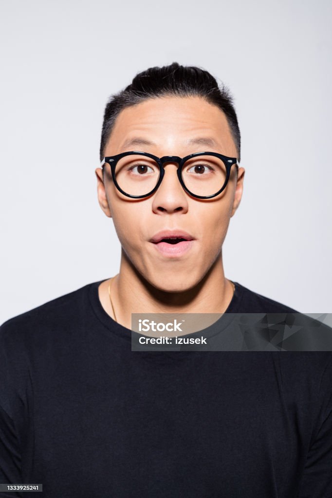 Surprised asian young man in black clothes Headshot of asian young man wearing black t-shirt and eyeglasses, looking at camera. Studio shot, grey background. Men Stock Photo
