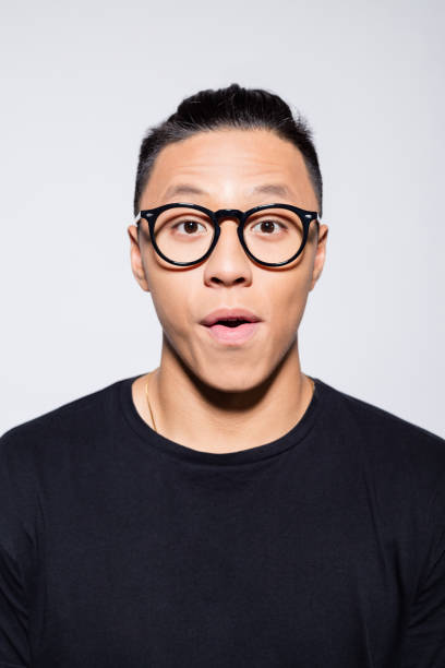 Surprised asian young man in black clothes Headshot of asian young man wearing black t-shirt and eyeglasses, looking at camera. Studio shot, grey background. confusion raised eyebrows human face men stock pictures, royalty-free photos & images