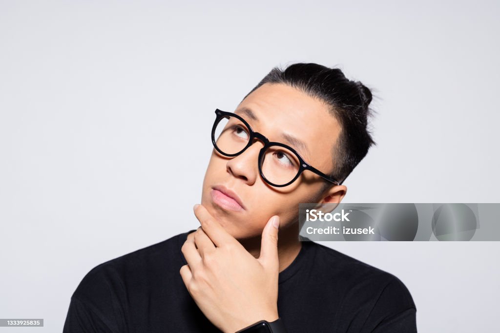 Thoughtful asian young man in black clothes Headshot of pensive asian young man wearing black t-shirt and eyeglasses, looking away with hand on chin. Studio shot, grey background. 20-24 Years Stock Photo