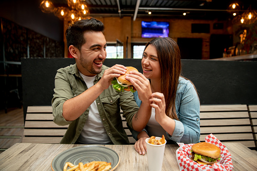 Very happy couple on a date at a restaurant eating hamburgers and man sharing his burger with his girlfriend - fast food concepts