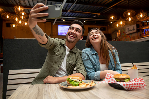Happy Latin American couple taking a selfie while eating hamburgers at a fast food restaurant - lifestyle concepts