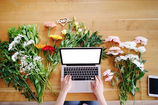 Woman hands typing on keyboard on a laptop with white screen surrounded by colorful fresh flowers and inscription love is. Valentine day concept