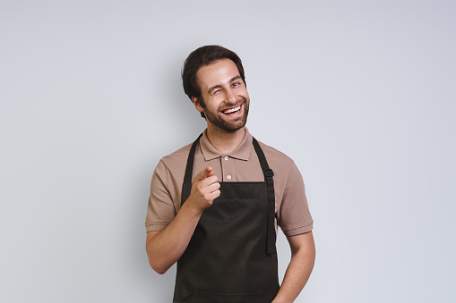Handsome young man in apron looking at camera and pointing you while standing against gray background
